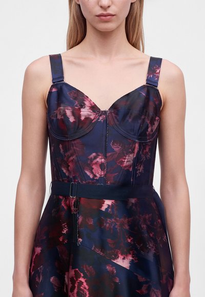 Femme portant une robe sans manches à imprimé floral foncé, avec un corsage ajusté, des bonnets structurés, des bretelles réglables et une ceinture assortie à la taille.