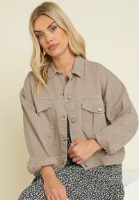 Beige denimjacka med klassisk krage, två bröstfickor och knappstängningar; har en avslappnad passform och uppvikta ärmar.