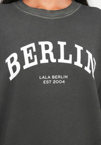 Γκρί φούτερ με μεγάλο λευκό κείμενο που γράφει "ΒΕΡΟΛΙΝΟ" πάνω από μικρότερο κείμενο "LALA BERLIN EST 2004." Ραμμένο λαιμό και μαλακή υφή.