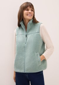 Ljusblå fleeceväst med hög krage, dragkedja och sidfickor; lagd över en krämfärgad långärmad topp med mjuk textur.