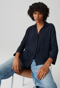 Camicia a maniche lunghe blu navy con colletto, tessuto morbido, vestibilità rilassata e chiusura con bottoni. Abbinata a jeans azzurri chiari.