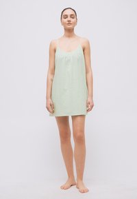 Νεαρή γυναίκα όρθια, ξυπόλυτη, φορώντας ένα ανοιχτοπράσινο αμάνικο κοντό φόρεμα, σε απλό λευκό φόντο.