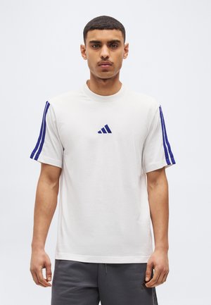 Άνδρας που στέκεται προς τα εμπρός φορώντας λευκή μπλούζα με μπλε ρίγες στα μανίκια και ένα μικρό μπλε λογότυπο στο κέντρο.