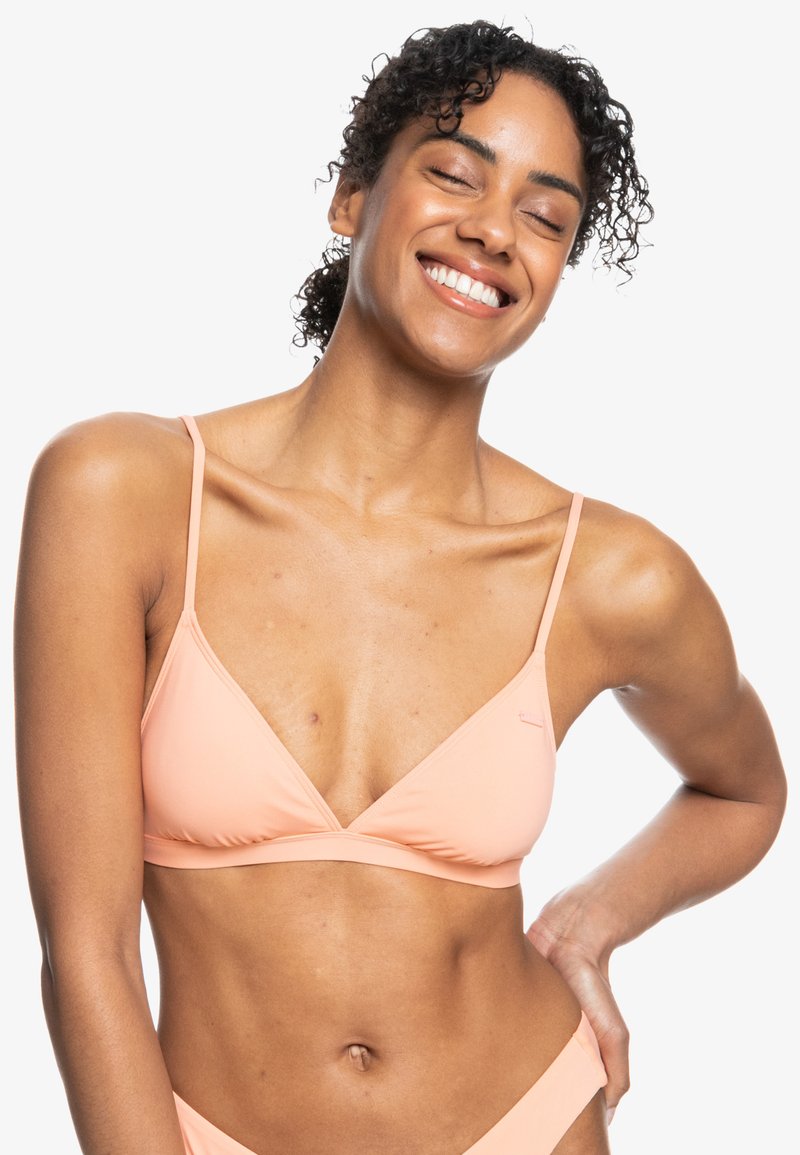 Perzikdriehoekbikini-top met dunne bandjes, zachte textuur en een kleine logo-accent. Model glimlachend, toont het ontwerp van de zwemkleding.