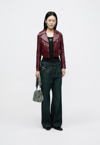 Bordeauxroter, kurz geschnittene Lederjacke, schwarzes Tanktop, dunkle Jeans mit weitem Bein und grüne, strukturierte Handtasche. Minimalistisches Design mit silberfarbenen Hardware-Akzenten.