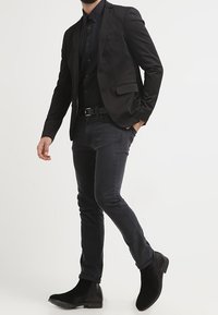 Man in een zwart colbert, zwart shirt, donkergrijze jeans en zwarte laarzen, lopend met één hand in zijn zak tegen een witte achtergrond.