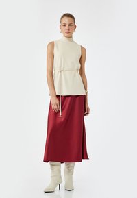 Beige mouwloos topje met hoge hals, vastgeknoopt in de taille, gecombineerd met een rode satijnen maxi rok en crèmekleurige kniehoge laarzen. Soepen texturen.