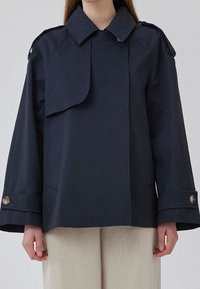 Navy blue cropped rain jacket with a large collar, wide sleeves, side pockets, and buttoned cuffs, made from smooth, water-resistant material.