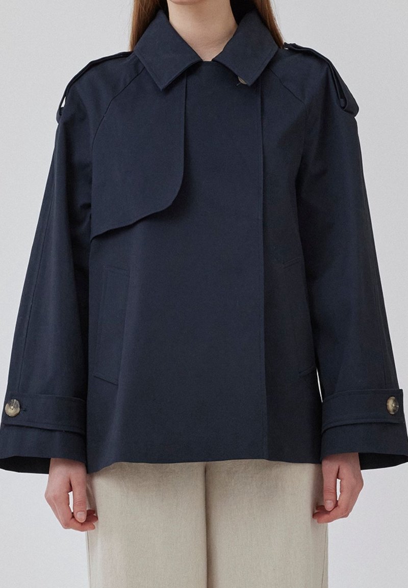 Navy blue cropped rain jacket with a large collar, wide sleeves, side pockets, and buttoned cuffs, made from smooth, water-resistant material.