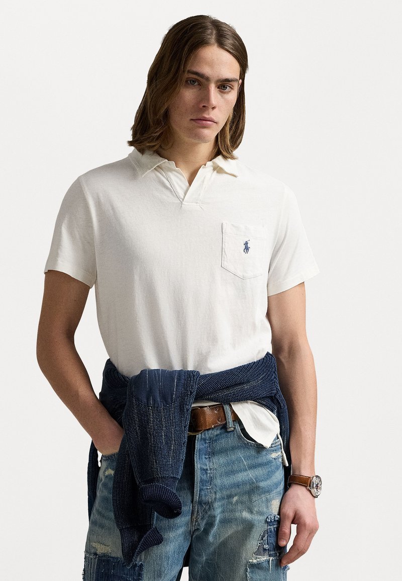 Beige polo shirt with a collar and a pocket, paired with distressed denim shorts. A dark blue knit sweater is tied around the waist.