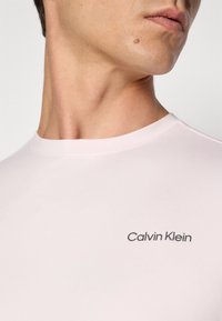 Roza kratka majica s kratkimi rokavi iz gladkega bombaža, ki ima črni logotip "Calvin Klein" na levem prsnem predelu in okroglo ovratnico.