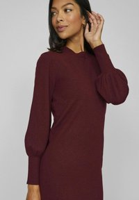 Burgundy klänning i stickad mjuk kvalitet med hög krage, ribbade mudd och puffärmar. Slät textur med figurnära silhuett.