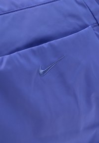 Κοντινό πλάνο μωβ υφάσματος με κεντημένο λογότυπο Nike Swoosh και οριζόντια λεπτομέρεια ραφής.