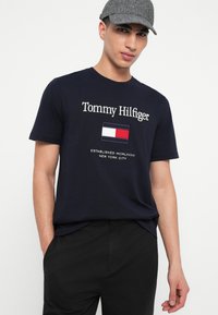 Άνδρας που φοράει γκρίζο καπέλο, μπλε μπλούζα Tommy Hilfiger με λευκό, κόκκινο και μπλε λογότυπο, και μαύρο παντελόνι, στέκεται μπροστά σε άσπρο φόντο.