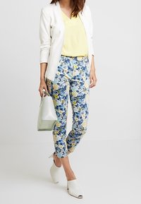 Vit blazer, gul V-ringad topp, byxor med blommönster i blått, gult och vitt; grön handväska; vita sandaletter med öppen tå.