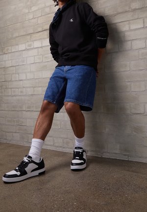 Person iført sort Calvin Klein sweatshirt, blå denimshorts, hvide mellemlange sokker og sorte og hvide sneakers, der står op ad en murstensvæg.