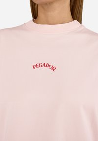 Rožinė medvilninė marškinė su raukšlėtu apykaklės dizainu ir raudonu išlenktu "PEGADOR." logotipu ant krūtinės. Minkšta tekstūra su laisvu kirpimu.