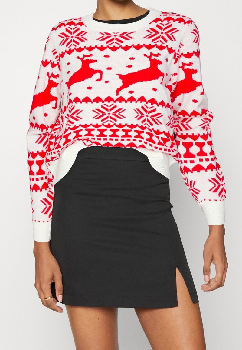Pull de Noël blanc et rouge avec motifs de rennes et flocons de neige, accompagné d'une mini-jupe noire avec une petite fente latérale.