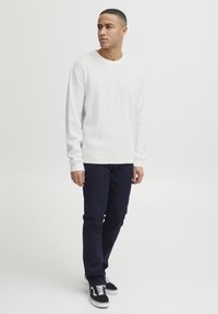 Chemise blanche à manches longues texturée, col rond, associée à un jean foncé et des baskets noires avec des lacets blancs. Design simple et moderne.