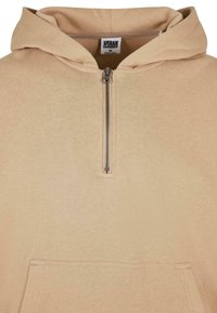 Sweat à capuche beige à fermeture éclair quart de tour en métal, poche kangourou à l'avant, et étiquette de marque "Urban Classics" à l'intérieur du col.