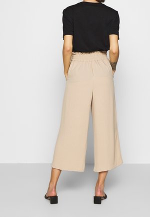 Persoon staat met de rug naar voren, draagt een zwarte top met korte mouwen, beige wijde cropped broek met elastische taille en zwarte sandalen met hak.
