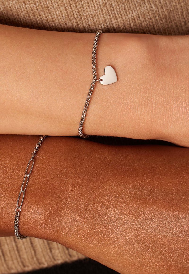 Twee armbanden om verschillende polsen: de ene met een klein zilveren hartje als bedel en een gestructureerde ketting, de andere met een gladde schakelketting.