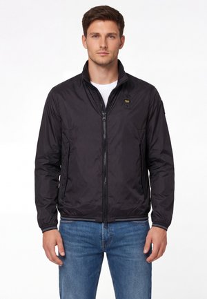 Uomo visto di fronte che indossa una giacca nera con zip sopra una maglietta bianca e jeans blu su uno sfondo bianco.