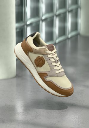 Baskets décontractées beige et marron avec lacets et semelle texturée, flottant au-dessus d'un sol gris réfléchissant près d'un mur en briques de verre.