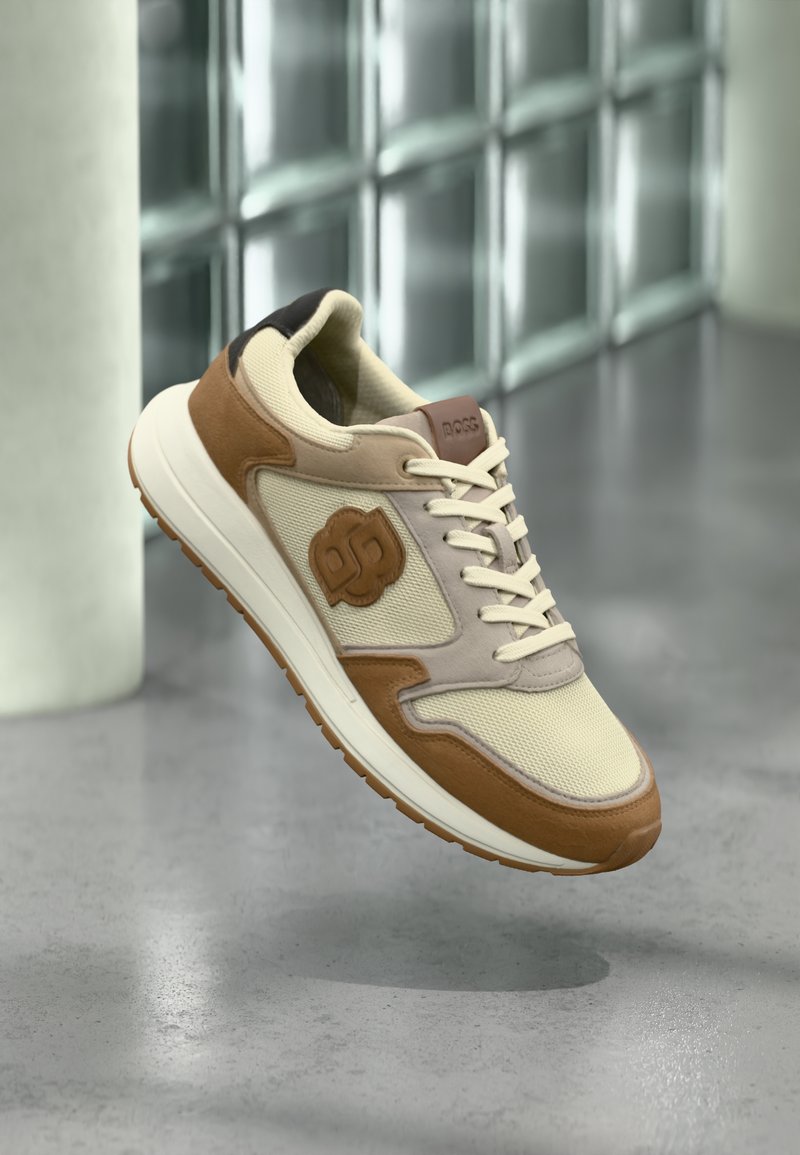 Baskets décontractées beige et marron avec lacets et semelle texturée, flottant au-dessus d'un sol gris réfléchissant près d'un mur en briques de verre.