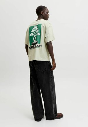 Personne portant un T-shirt beige oversize avec un motif bonsaï vert et un texte au dos, associé à un jean noir ample et des chaussures marron, debout de côté.