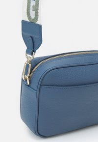 Blå läder crossbody-väska med guldrippar, med en texturerad yta och ett tygband med grönt tryck. Kompakt rektangulär form.