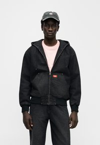 Μαύρο τζιν φούτερ με φερμουάρ, μπροστινή τσέπη καγκουρό, ρυθμιζόμενο κορδόνι στο καπό και κόκκινη ετικέτα. Φορεμένο πάνω από ένα ανοιχτό ροζ πουκάμισο.