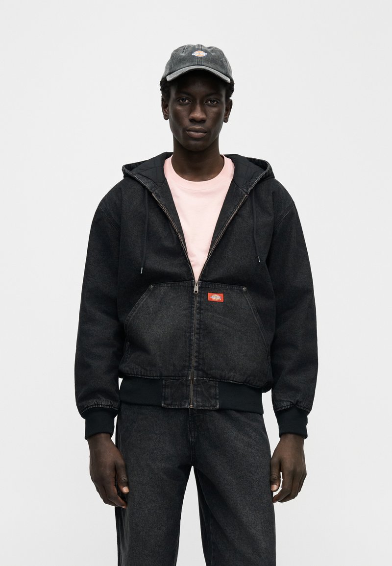 Zwarte denim capuchontrui met ritssluiting, een kangoeroezak aan de voorkant, verstelbare capuchon met trekkoorden en een rood label. Draagt een lichtroze shirt eronder.