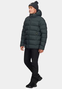 Ciemnozielona kurtka puchowa z kapturem, zapinana na zamek, z poziomymi przeszyciami. Nosi ją w połączeniu z czarnymi spodniami i solidnymi czarnymi butami.