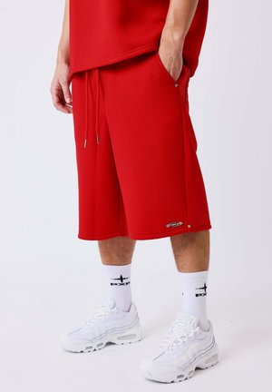 Persona con pantalones cortos rojos hasta la rodilla, calcetines blancos con logo negro y zapatillas deportivas blancas sobre un fondo claro y liso.