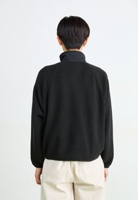 Persona con el cabello corto y oscuro, vistiendo una chaqueta de forro polar negra y pantalones beige claros, de espaldas contra un fondo blanco liso.