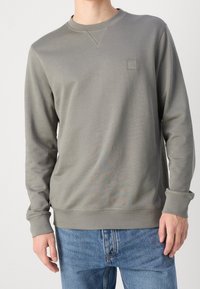 Man med grå långärmad sweatshirt med en liten "BOSS"-logotyp på bröstet och blå denimjeans, stående mot en vit bakgrund.