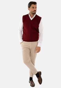 Gilet lavorato a maglia bordeaux sopra una camicia bianca, abbinato a pantaloni beige e mocassini marroni. Presenta un design a V, evidenziando una texture liscia.