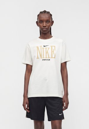Person iført en hvid Nike Sports Club T-shirt og sorte Nike shorts, står mod en ensartet lys baggrund.