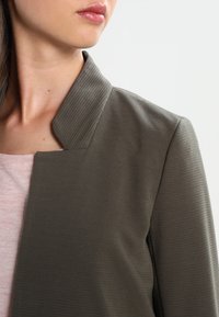 Veste texturée vert olive avec un col côtelé et un design à devant ouvert, portée sur un haut rose clair à motif subtil.