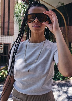 Młoda kobieta z długimi warkoczami poprawia duże czarne okulary przeciwsłoneczne, ubrana w jasnoszarą koszulę i złote naszyjniki, stoi na świeżym powietrzu w świetle słonecznym.