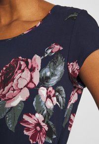 Top floreale blu navy con grandi rose rosa e foglie verdi, maniche corte a cappuccio e una texture liscia. Collo con accenti floreali.