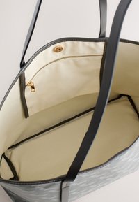 Sac fourre-tout ouvert avec intérieur en tissu beige, anses noires et une poche intérieure zippée avec fermeture éclair dorée et bouton-pression.