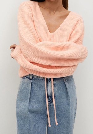 Femme portant un pull en maille rose doux col en V avec de longues manches surdimensionnées, associé à un jean bleu ample taille haute avec un cordon rose.