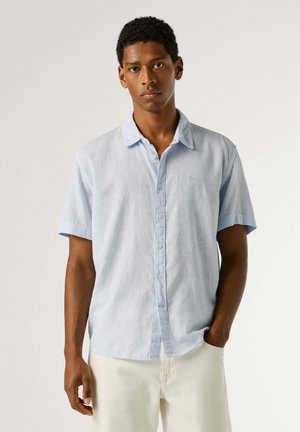 Noor mees, kes kannab helehalli lühikeste varrukatega nööpidega särki ja helevalgeid pükse, seisab ühe käega taskus.