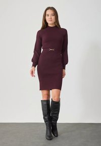 Eine Frau trägt ein enganliegendes, langärmeliges burgunderfarbenes Strickkleid mit einem dünnen Gürtel und schwarzen kniehohen Stiefeln. Sie steht auf einem grauen Boden vor einer weißen Wand.