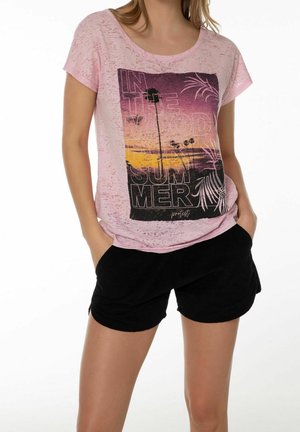 Kobieta w jasnoróżowej koszulce z nadrukiem zachodu słońca i palmy oraz czarnych szortach, ręce w kieszeniach.