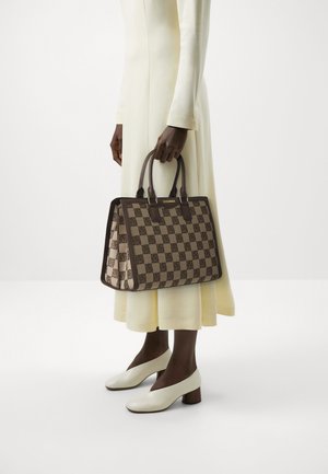 Persona con vestido largo color crema sostiene un bolso tote marrón con estampado a cuadros y asas oscuras, llevando zapatos blancos de tacón cuadrado, sobre fondo blanco liso.