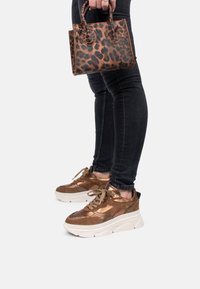 Zapatillas metálicas bronce con suelas gruesas blancas, combinadas con un pequeño bolso marrón de estampado leopardo. Unos jeans skinny negros complementan el conjunto.