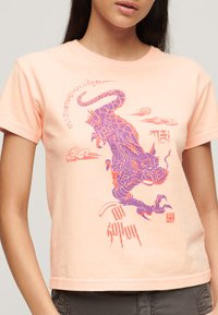 Camiseta corta de algodón en color melocotón con un gráfico de dragón púrpura con acentos rojos y texto estilizado, mangas cortas y cuello redondo.